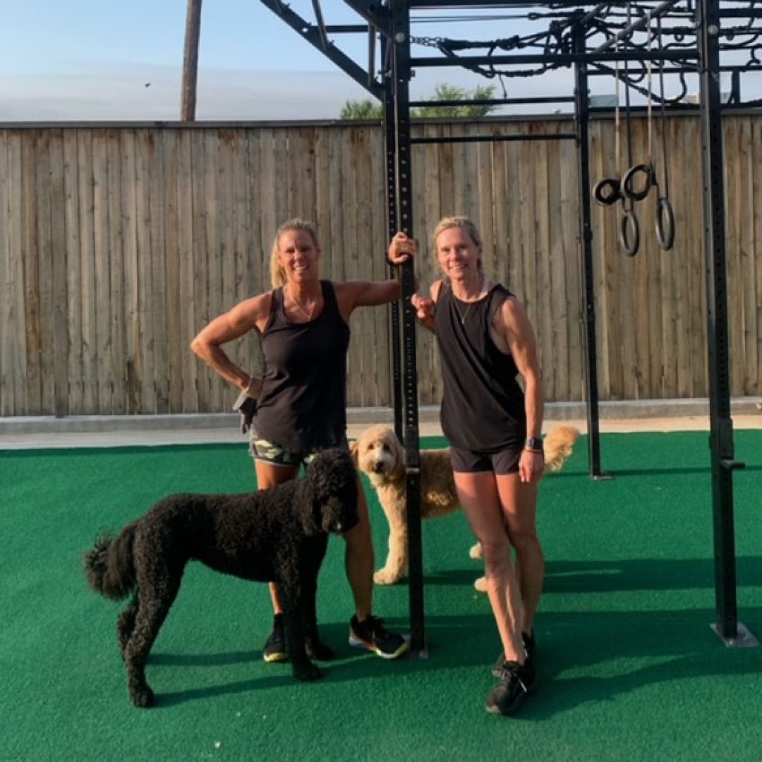 Coach Amanda and Coach Stacy in the outdoor part of HIIT4FIT surrounded by gym equipment and two adorable dogs